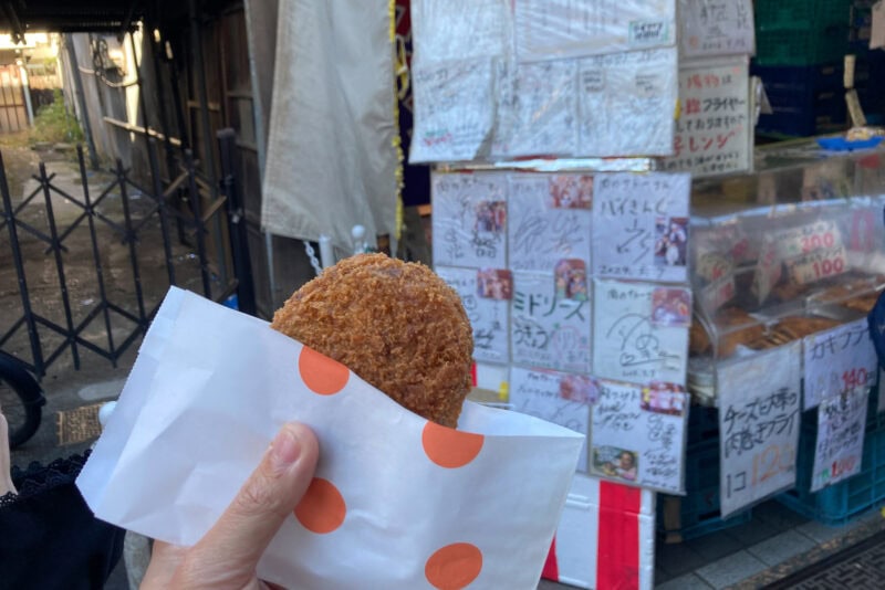 A hand holds a fried snack in a white paper bag with orange dots, in front of a shop on Yanaka Ginza, where menus and posters are displayed on the wall outside.