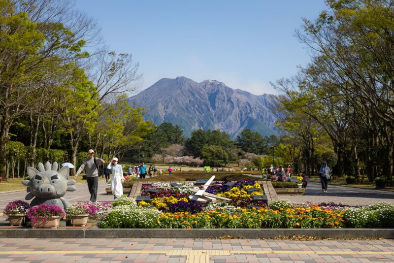 Yoshino Park in Kagoshima offers a scenic escape with colorful flower beds, a charming cow statue, and walking paths set against a tall, forested mountain under a clear blue sky.