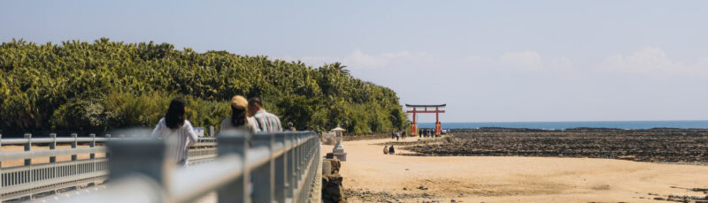 People walk along a bridge toward a sandy beach with a red torii gate nearby, dense green trees on the left, and the ocean and rocky shore in the background under a clear sky.