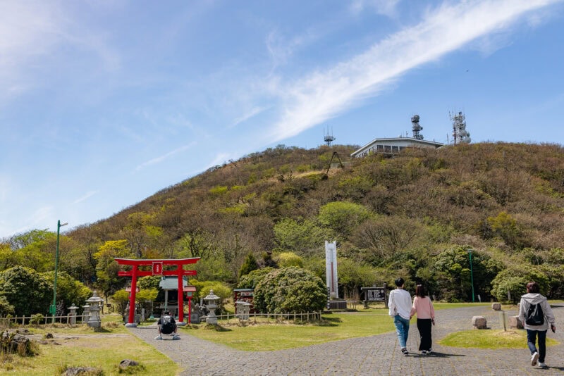People walk on a stone path toward a red torii gate and shrine at the base of a green hill, topped with communication towers and a building, under a blue sky with wispy clouds.