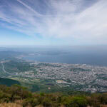 Beppu Ropeway: Ride to the Best Views in Beppu A wide aerial view of a coastal city bordered by greenery and mountains, with blue ocean and sky above. Wispy clouds stretch across the sky, and the cityscape extends toward the shoreline.