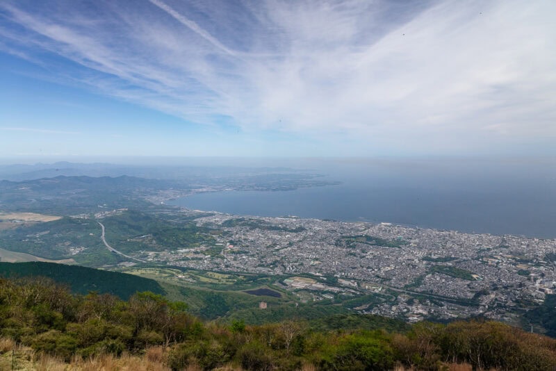 A wide aerial view of a coastal city bordered by greenery and mountains, with blue ocean and sky above. Wispy clouds stretch across the sky, and the cityscape extends toward the shoreline.
