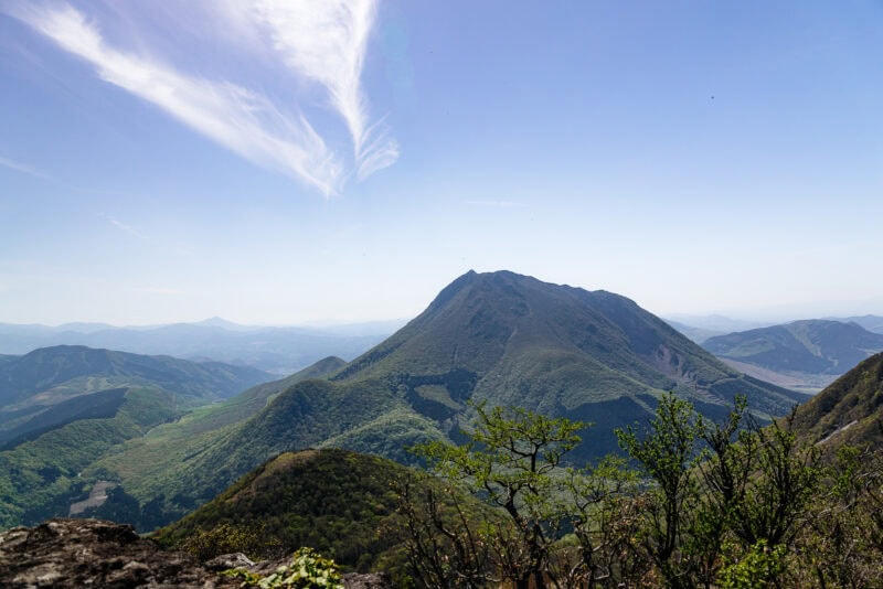 A distant, tree-covered mountain rises under a clear blue sky, with a wispy white cloud overhead and green hills rolling away into the horizon. Sparse vegetation is visible in the foreground.