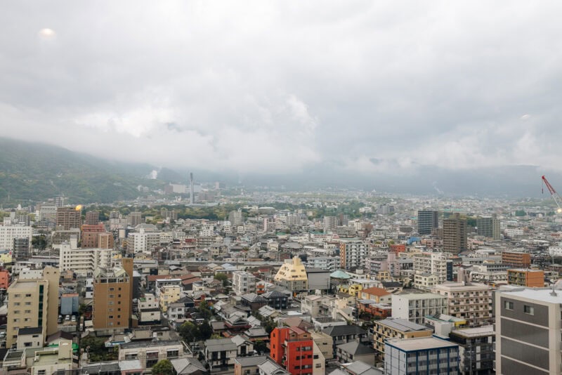 A wide view of a city with numerous buildings and houses, surrounded by misty mountains under a cloudy, overcast sky. The cityscape is densely packed and stretches towards the horizon.