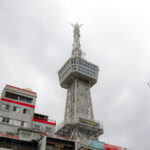 A tall, silver observation tower with a lattice structure rises above nearby buildings under a cloudy sky. The tower has an enclosed viewing platform near the top.