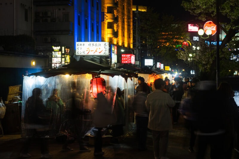 A bustling street at night in Japan with people gathered around a brightly-lit outdoor ramen stall, illuminated by red lanterns and surrounded by colorful neon signs.