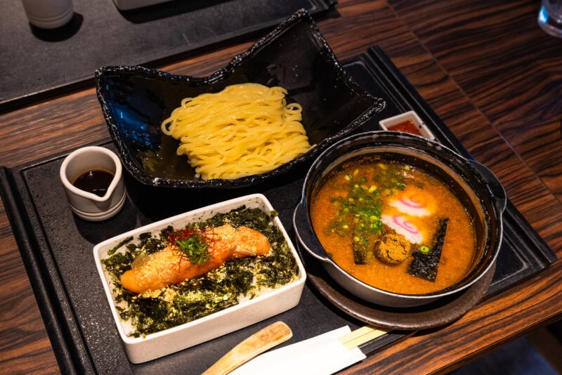 A Japanese meal set on a tray with noodles, a bowl of broth topped with green onions and fish cake, and a side dish of rice garnished with seaweed and a piece of grilled salmon.