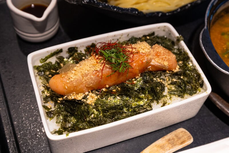 Rectangular white dish filled with steamed rice, topped with seasoned seaweed, a grilled sausage, chopped herbs, sesame seeds, and thin red pepper strands. A small jug of sauce is in the background.