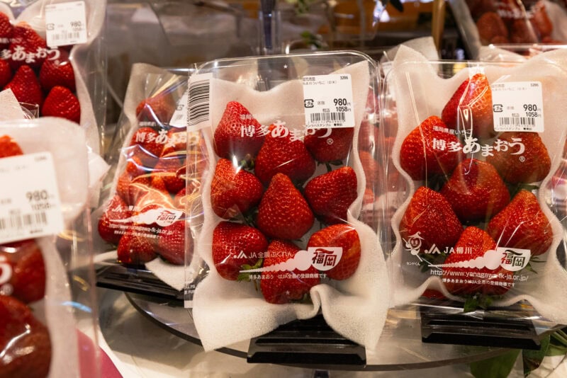 Plastic containers of large, fresh strawberries with Japanese labels and price tags are displayed for sale in a store. The strawberries are arranged neatly on white foam trays inside the packaging.