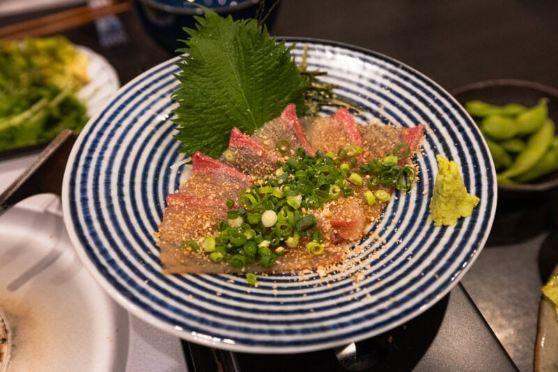 Thin slices of sashimi are arranged on a striped plate, garnished with chopped green onions, sesame seeds, and accompanied by a shiso leaf and a small mound of wasabi on the side.