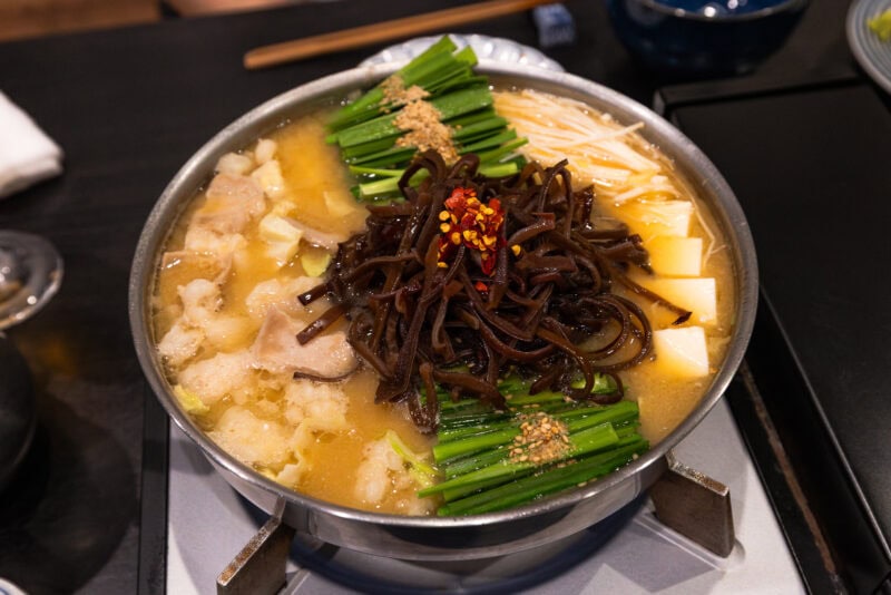 A hot pot filled with broth, sliced meat, tofu, green onions, enoki mushrooms, wood ear mushrooms, chili flakes, and sesame seeds on a table, ready to be served.