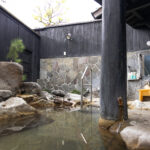 Hyotan Onsen: Beppu's Famous Hot Spring An outdoor Japanese onsen bath with natural rocks, clear water, and a stone wall. Two yellow stools sit in front of a mirror and toiletries on the right, with greenery and wooden walls surrounding the area.