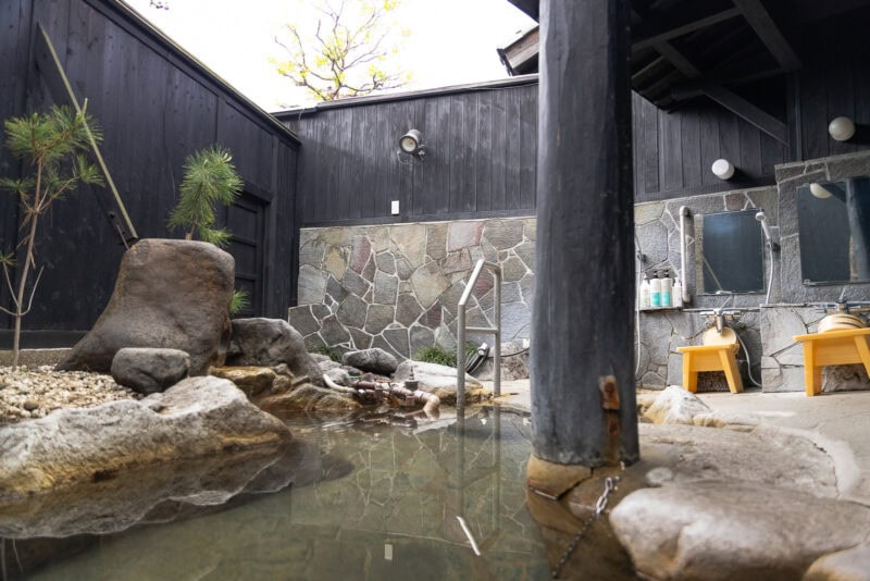 An outdoor Japanese onsen bath with natural rocks, clear water, and a stone wall. Two yellow stools sit in front of a mirror and toiletries on the right, with greenery and wooden walls surrounding the area.