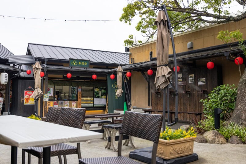 An outdoor seating area with tables and chairs in front of a small restaurant decorated with red lanterns, plants, and string lights. The scene is calm and inviting, with umbrellas closed above some tables.