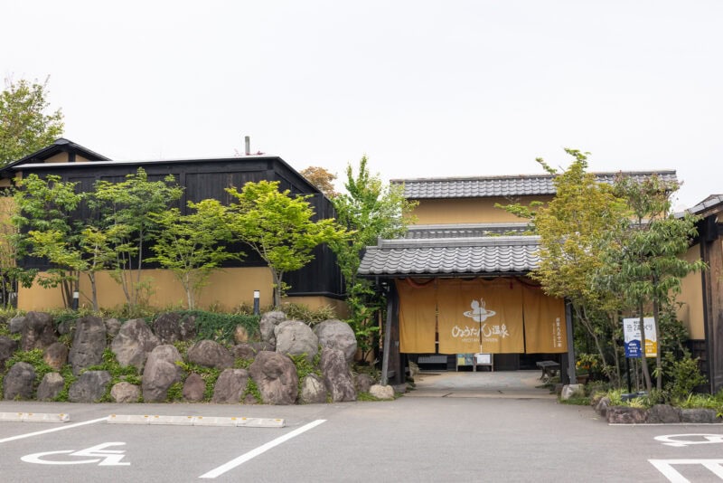A traditional Japanese building with a tiled roof, tan walls, and a large fabric sign at the entrance, surrounded by trees and rocks, with parking spaces in the foreground.