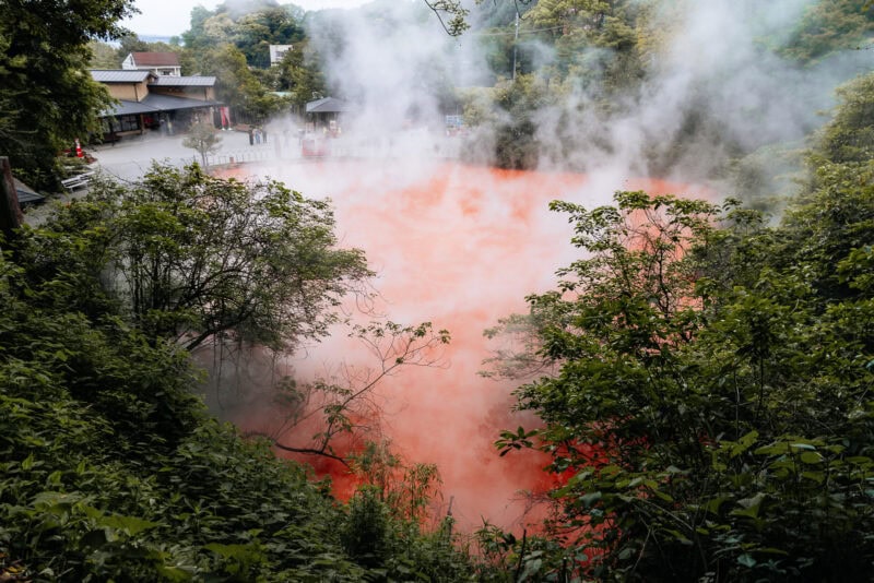 A hot spring with red-colored water emits thick steam, surrounded by dense green foliage. Buildings are visible in the background, and the scene appears to be in a natural park or tourist area.