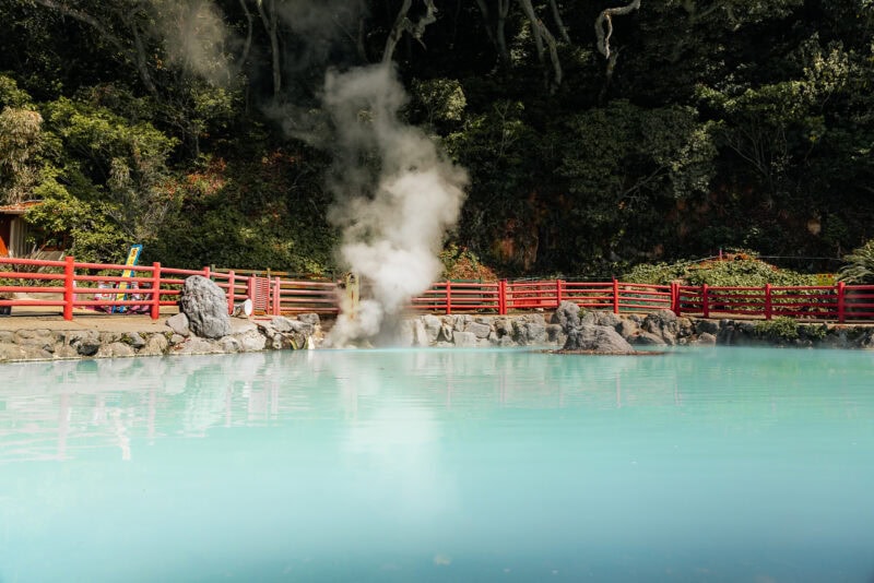 A steaming hot spring pool with pale blue water, surrounded by rocks and a bright red fence, with lush green trees in the background.