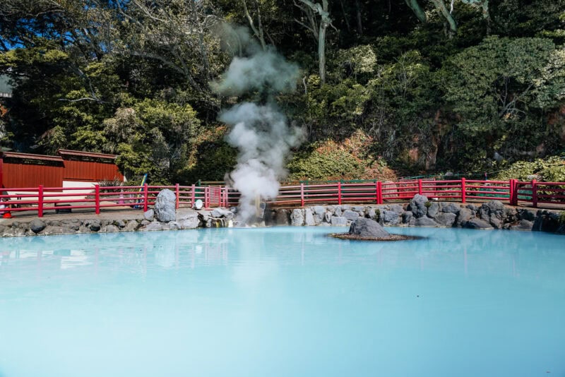 A steaming hot spring with milky blue water is surrounded by rocks, red wooden fences, and lush green trees in the background. Steam rises from the center of the pond.
