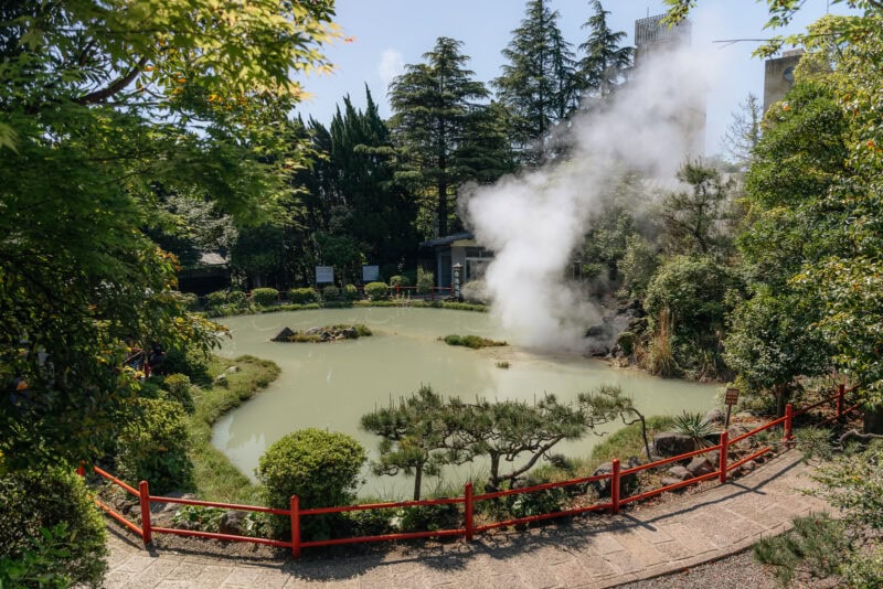 A round, milky-green pond surrounded by lush trees and shrubs, with steam rising from the water, and a red fence encircling the pond on a sunny day.