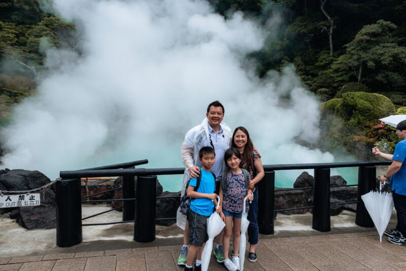A family of four poses and smiles in front of a steaming hot spring with lush greenery in the background. The parents stand behind their two children, all wearing casual clothing. A black fence separates them from the steam.