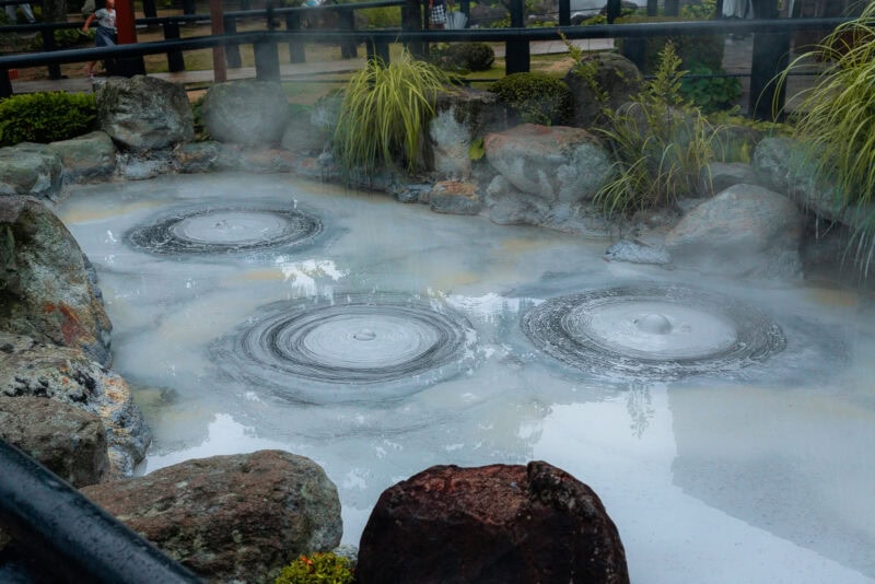 Three round, bubbling pools of milky water surrounded by rocks and plants, with steam rising into the air, likely at a hot spring or geothermal area in a garden setting.