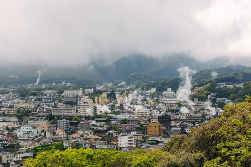 A cityscape with numerous buildings and houses, with steam rising from several spots. Misty mountains loom in the background under a cloudy sky, and green foliage is visible in the foreground.