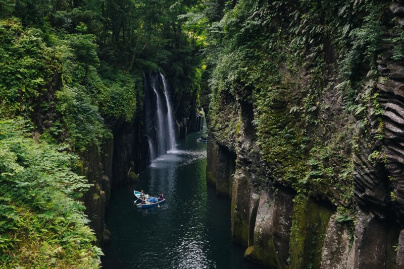 A narrow river gorge filled with lush green vegetation, featuring steep rocky cliffs and a waterfall. People in small blue boats paddle along the calm, dark water near the base of the waterfall.