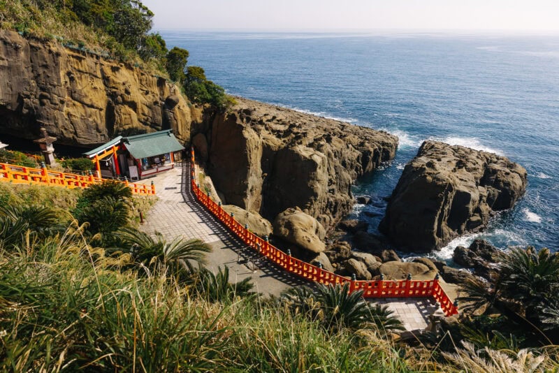 A coastal shrine with a green roof sits among rocky cliffs by the sea, connected by a path lined with bright red railings. Waves crash against the rocks, and lush greenery surrounds the area.