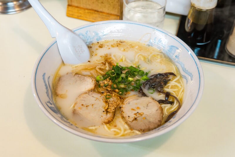 A bowl of ramen with creamy broth, sliced pork, noodles, green onions, mushrooms, and spices, served with a white soup spoon on a light-colored table.