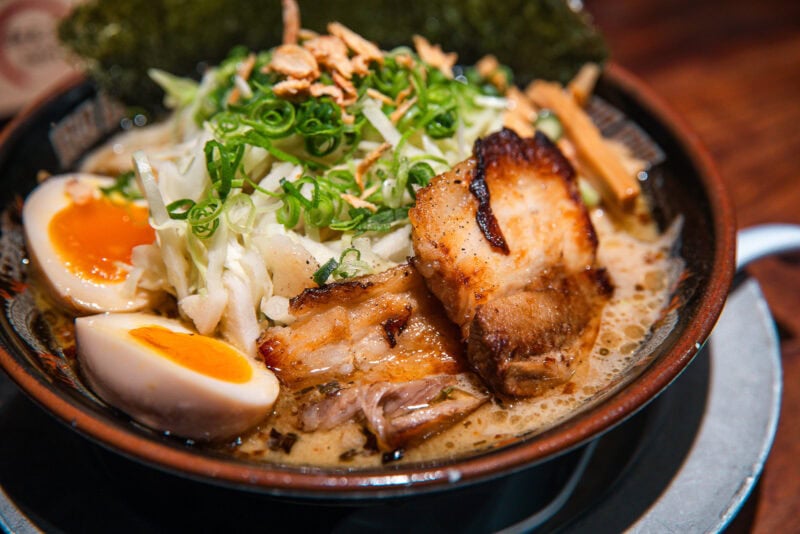 A bowl of ramen with sliced pork, half a soft-boiled egg, chopped green onions, shredded cabbage, bamboo shoots, and seaweed in a rich broth.