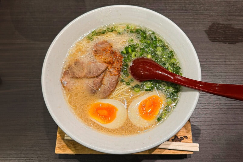 A bowl of ramen topped with two halves of soft-boiled eggs, sliced pork, chopped green onions, and a red soup spoon, placed on a wooden table with chopsticks resting underneath the bowl.
