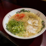 Kyushu Ramen: The Ultimate Guide to Kyushu's Famous Noodles A bowl of ramen with sliced pork, chopped green onions, red pickled ginger, and sesame seeds in a clear broth, served on a red table with wooden chopsticks beside it.