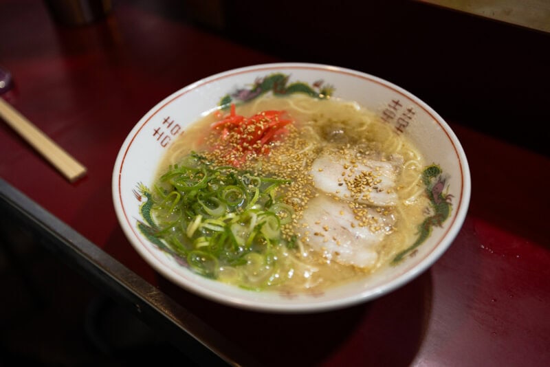 A bowl of ramen with sliced pork, chopped green onions, red pickled ginger, and sesame seeds in a clear broth, served on a red table with wooden chopsticks beside it.