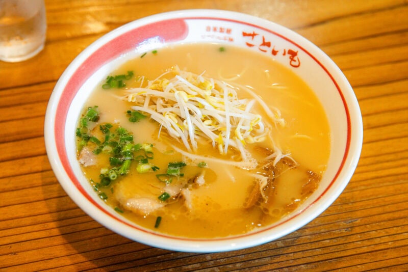 A bowl of ramen with creamy broth, topped with bean sprouts, chopped green onions, and slices of meat, served in a white bowl with red trim on a wooden table.