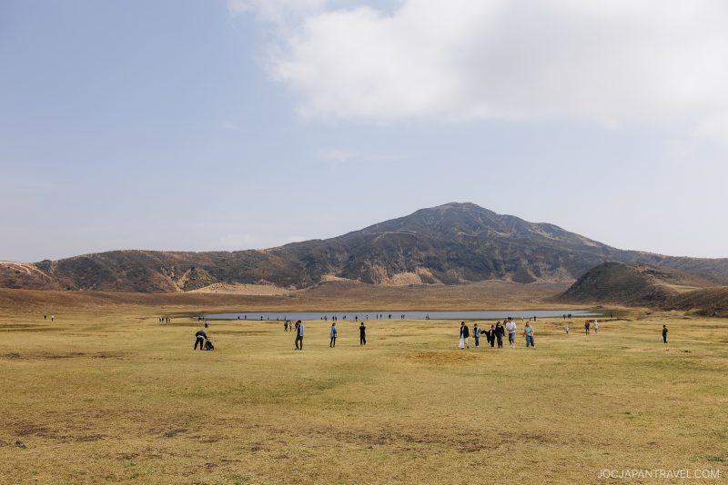 People walk and relax on a wide grassy plain with a shallow pond in the distance, backed by a large mountain under a partly cloudy sky.