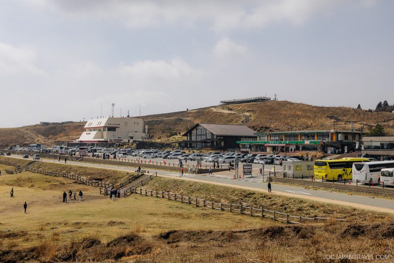 A wide view of a parking area with several buildings, cars, and buses near a grassy field. People walk along a path with wooden fences, and the background features rolling hills under a partly cloudy sky.