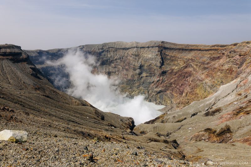 Crater of a volcanic mountain with steep, rocky walls and white steam or gas rising from within; the landscape appears barren and rugged under a hazy sky.