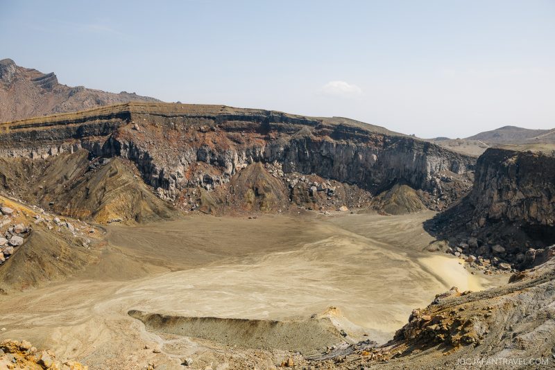 A wide view of an empty volcanic crater with rocky, steep walls and a flat, sandy floor under a clear blue sky. Sparse vegetation is visible on the surrounding slopes.