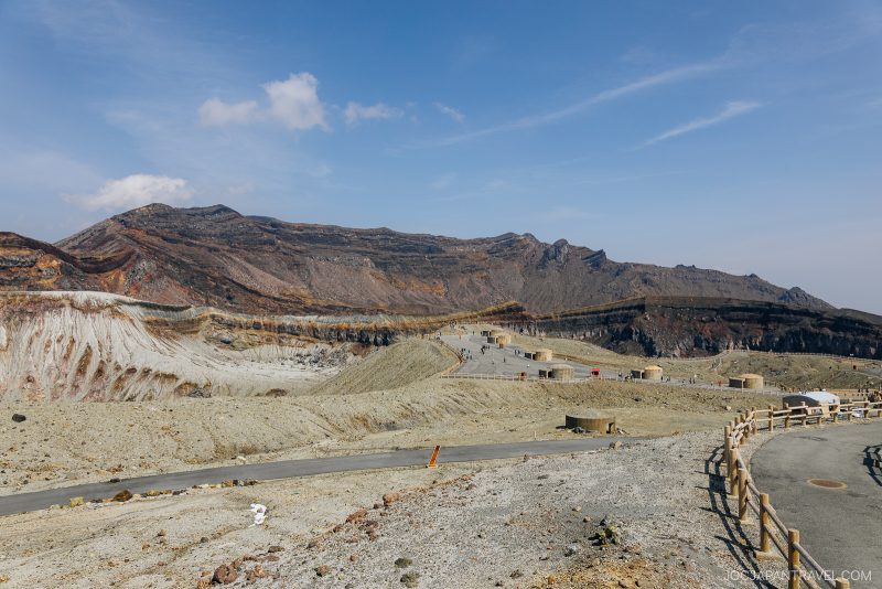 A wide view of Mount Aso’s volcanic crater in Japan, showing rugged, rocky terrain with a pathway, fences, and small buildings under a clear blue sky.