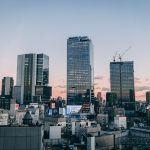 How to Survive Summer in Japan: Essential Tips for Heat, Humidity, and Travel A cityscape at sunset shows modern high-rise buildings and construction cranes against a clear sky, with smaller buildings in the foreground and a soft pink and blue horizon.