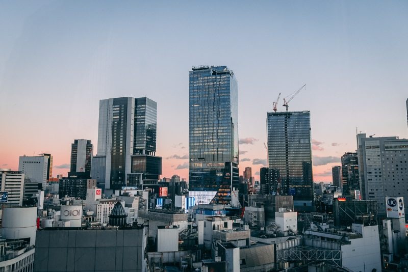 A cityscape at sunset shows modern high-rise buildings and construction cranes against a clear sky, with smaller buildings in the foreground and a soft pink and blue horizon.