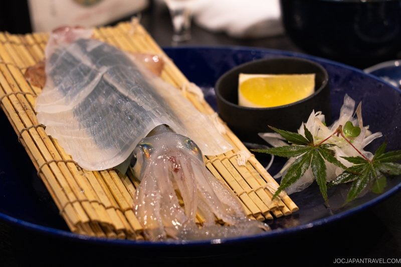 A plate features a translucent, raw squid served on a bamboo mat with lemon, garnish, and thinly sliced sashimi, presented on a dark blue dish.