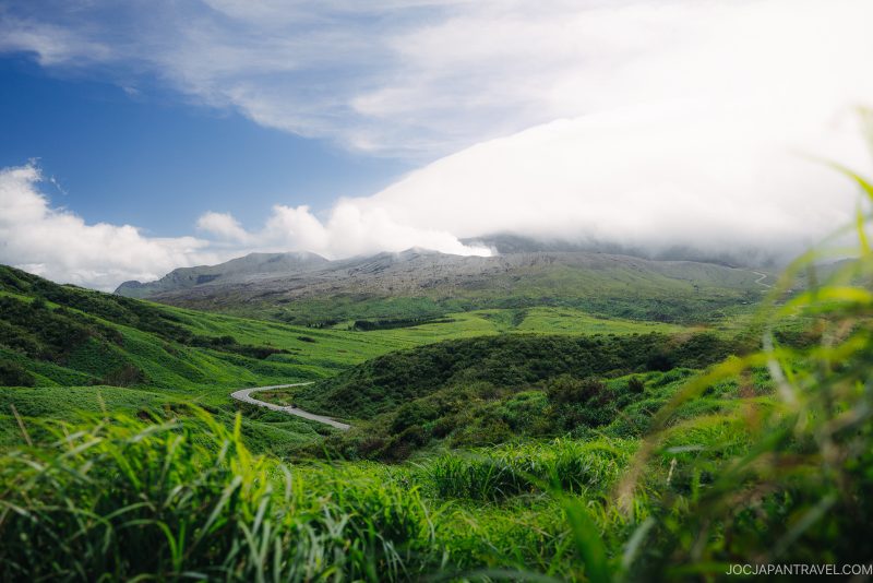 A winding road cuts through lush green hills under a bright blue sky with scattered clouds, and a mountain partially covered by mist in the background.