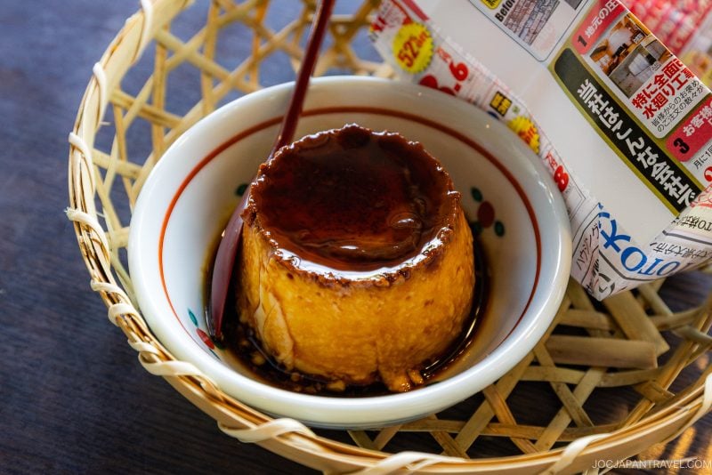 A bowl of creamy caramel flan is placed in a woven basket, with caramel sauce being poured on top. A partially visible Japanese newspaper is beside the bowl.
