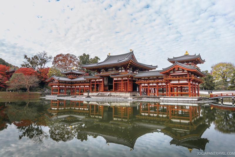 Byodoin Temple in Uji, Kyoto, with its striking red wooden architecture, is beautifully reflected in a calm pond and surrounded by autumn trees under a partly cloudy sky.
