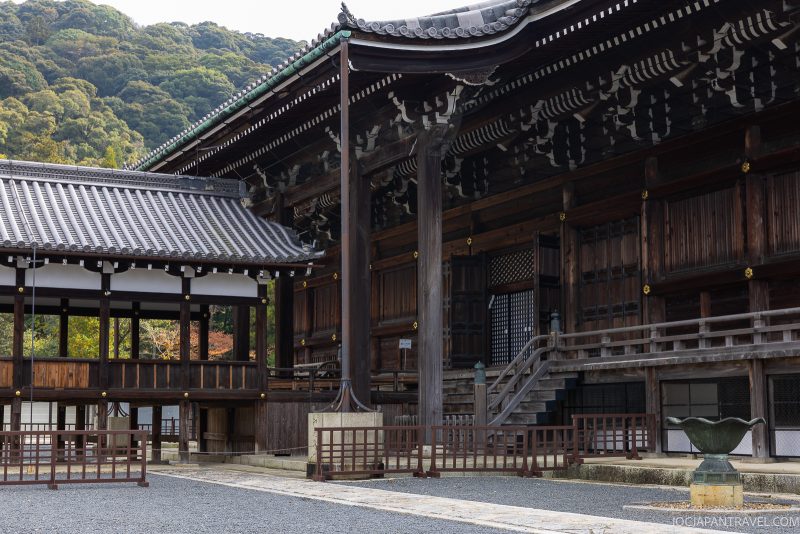 A traditional wooden Japanese temple with tiled roofs stands beside gravel paths and a low fence, surrounded by greenery and the forested hills of Higashiyama. This picturesque scene evokes the timeless beauty found at Kyoto temples such as Chionin.