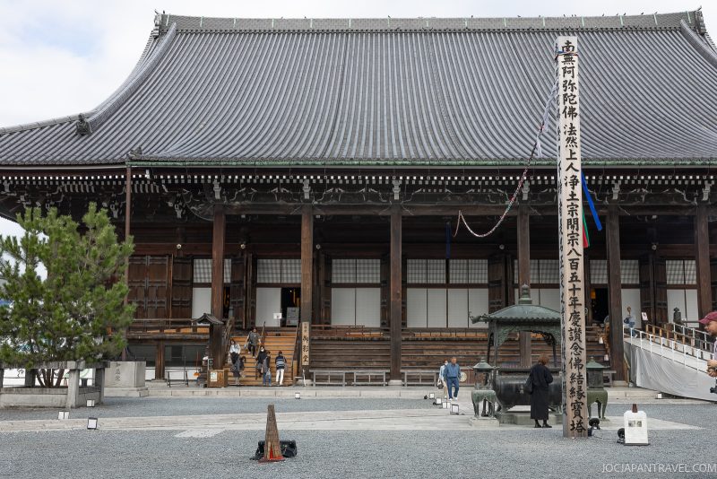 Large traditional Japanese temple with a sloped tiled roof, wooden pillars, and a tall wooden signpost with Japanese writing. People are walking and standing in front of the Chionin temple gate in Kyoto on a gravel courtyard.