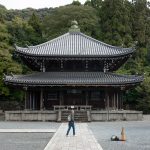Chionin Guide: Japan's Largest Temple Gate and Bell in Kyoto's Higashiyama A person walks toward the traditional Chionin temple gate in Kyoto, surrounded by trees, with a stone path leading to the entrance and a green incense burner on the left.