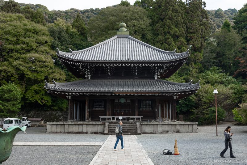 A person walks toward the traditional Chionin temple gate in Kyoto, surrounded by trees, with a stone path leading to the entrance and a green incense burner on the left.