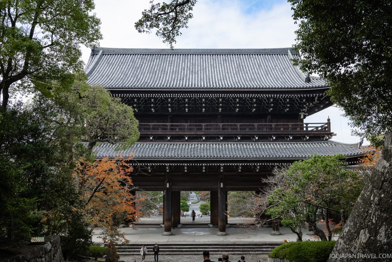 A large traditional Japanese temple gate with a tiled roof, surrounded by green trees and a few people walking underneath. Stairs lead up to the impressive Chionin temple gate in Kyoto, and autumn-colored leaves are visible on some trees.